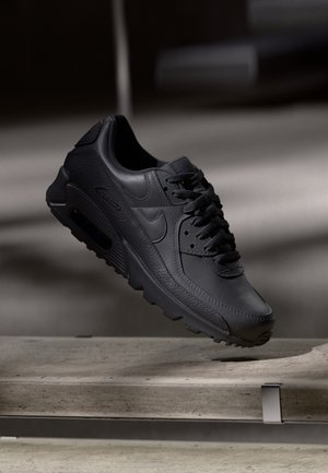 Sort Nike Air Max sneaker svævende over en betonbænk i et svagt oplyst indendørs miljø.