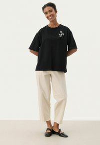 Camiseta de algodón negra con mangas cortas y bordado floral en el lado izquierdo del pecho, combinada con pantalones beige claros ajustados y zapatos negros.