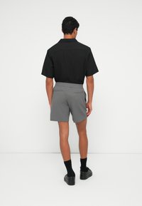 Black short-sleeve shirt with a pointed collar, paired with gray textured shorts. Model wears black socks and shoes, standing against a plain background.