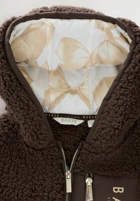 Giacca in pile marrone con una superficie morbida e testurizzata; presenta una chiusura con zip, una fodera interna a contrasto con motivi di fiocchi beige e un dettaglio con logo.