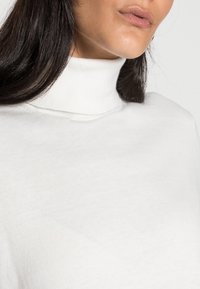 Pull à col roulé blanc avec une texture douce et lisse. Gros plan sur le col montant et la coupe décontractée, avec des détails de couture peu visibles.