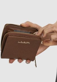 Portafoglio in pelle marrone con finitura texturizzata, chiusura con zip e accessori in tono oro; una mano tiene il portafoglio che mostra i compartimenti interni.