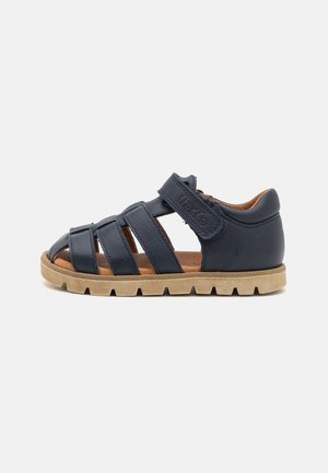Navy blue leather sandal with open toe and sides, featuring crisscross straps and adjustable Velcro closure. Light rubber sole with grooves.