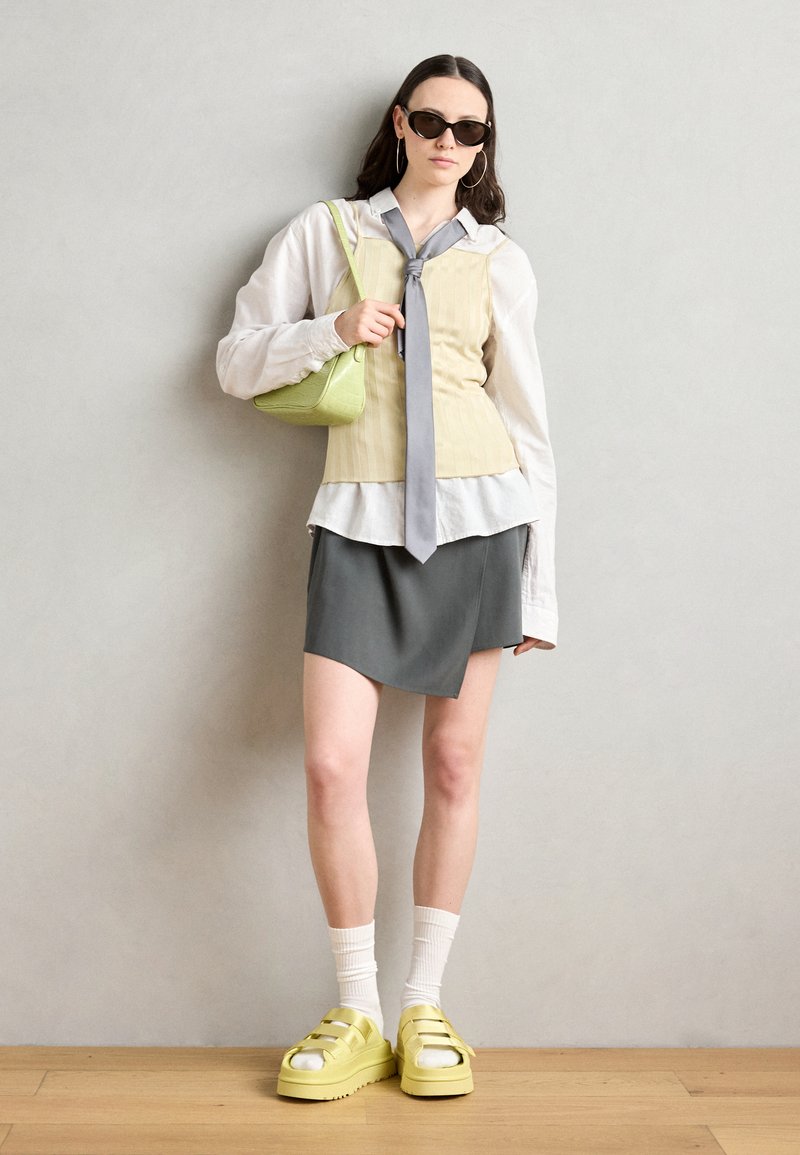 Top a coste giallo chiaro con un nodo grigio, camicia bianca sotto, gonna grigia asimmetrica e scarpe platform gialle chunky. Borsa verde e occhiali da sole.