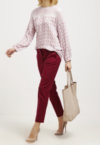Top a maniche lunghe rosa chiaro a fantasia con bordi in pizzo, abbinato a pantaloni aderenti bordeaux e una borsa a tracolla beige. Tacchi alti neutri completano il look.