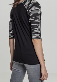 En person som bär en svart skjorta med grå och svart kamouflage i tre fjärdedelar ärmar och ljusgrå jeans, sedd bakifrån.