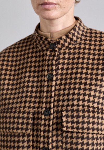 Bruine en tan houndstooth-gepatroneerde jas met hoge kraag, zwarte knopen en twee voorkruiden. Gelaagd stof, op maat gemaakte pasvorm.