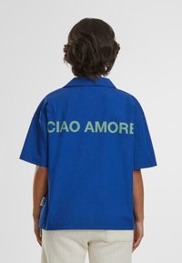 Blå kortærmet skjorte lavet af et glat stof har et ryggrafik med teksten "CIAO AMORE" i fed grøn skrift.