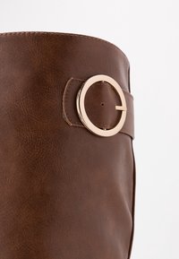 Botte en cuir synthétique marron avec un dessus arrondi, ornée d'une boucle circulaire dorée et d'une texture lisse. Détails de surpiqûres le long du bord.