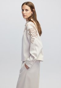 Lichtbeige sweatshirt met kantdetails op de schouders, zachte textuur, ontspannen pasvorm en lange mouwen. Gecombineerd met bijpassende lichtgekleurde broek.