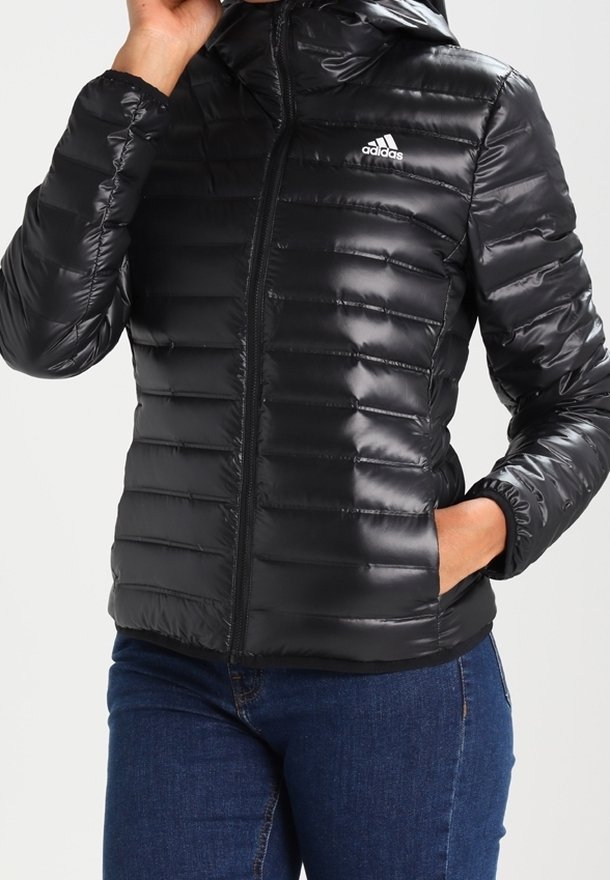 Femme portant une veste matelassée noire Adidas avec capuche, fermeture éclair à l'avant, une main dans la poche, associée à un jean bleu.