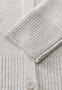 Lichtgrijze gebreide cardigan met een overlapping van de mouw en ribbelboord, voorzien van een enkele bijpassende knoop op een zachte stoftextuur.
