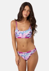 Femme aux longs cheveux foncés portant un soutien-gorge floral rose et des sous-vêtements assortis avec des bandes violettes marquées PSD sur fond blanc.