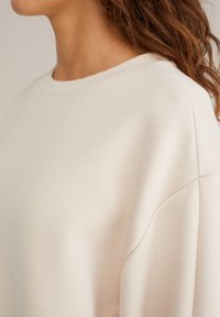 Primo piano di una persona che indossa una maglietta a maniche lunghe di color crema con scollatura rotonda e cucitura della spalla visibile.