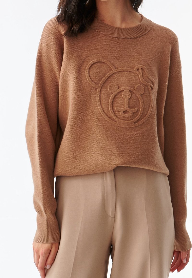 Pull marron avec un motif en relief de tête d'ours, porté avec un pantalon large beige clair sur une personne debout.