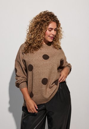 Kvinde med krøllet hår iført en brun sweater med mørke prikker og sorte bukser, der ser ned med et let smil og hånden i lommen.