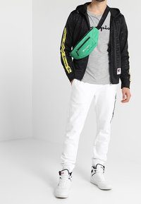 Svart zip-up hoodie med gula tryck, grå t-shirt, vita joggers och en grön axelväska; vita sneakers med svarta och röda detaljer.