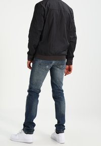 Schwarze Bomberjacke mit einem Saum und Bündchen, kombiniert mit blauen Jeans und weißen Sneakers. Glattes Material mit einer taillierten Silhouette.