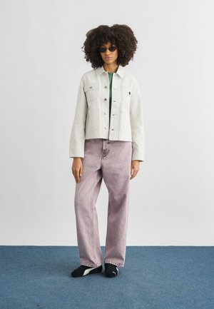 Kvinde med krøllet hår iført solbriller, en lysefarvet jakke, løstsiddende lavendelfarvede jeans og sorte sneakers, stående mod en ensfarvet baggrund.