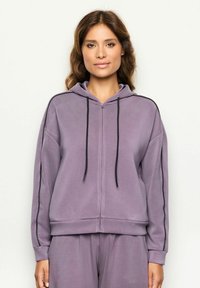 Mujer de piel clara con cabello castaño ondulado, vistiendo una sudadera con cremallera lila y pantalones a juego, contra un fondo simple.