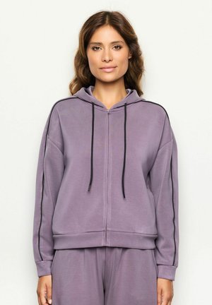 Mujer de piel clara con cabello castaño ondulado, vistiendo una sudadera con cremallera lila y pantalones a juego, contra un fondo simple.