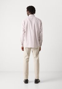 Camicia a maniche lunghe a righe in tessuto leggero, presenta sottili righe verticali rosse e bianche con una vestibilità rilassata. Abbinata a pantaloni beige e scarpe nere.