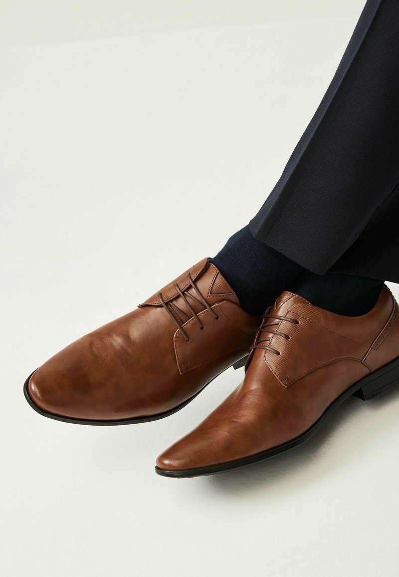 Brown leather shoes with a smooth finish, pointed toe, and lace-up design. Features a textured panel and black sole. Worn with navy socks.