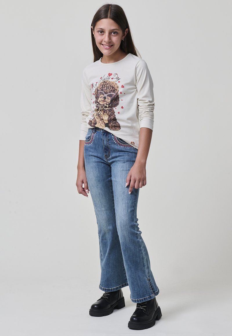 Ein junges Mädchen lächelt und trägt ein beiges Langarmshirt mit einem Hundemotiv, blaue Jeans mit mit Perlen besetzten Taschen und schwarze Plateaustiefel.