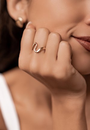 Goldfarbener Ring mit einem wellenförmigen Design, der an einer Hand getragen wird. Der Hautton ist hell, wodurch die reflektierende Oberfläche des Rings gut zur Geltung kommt.
