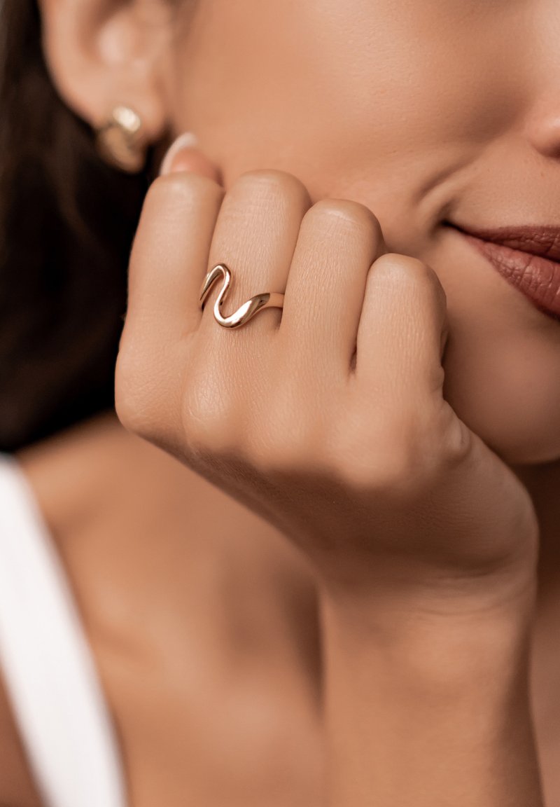 Goldfarbener Ring mit einem wellenförmigen Design, der an einer Hand getragen wird. Der Hautton ist hell, wodurch die reflektierende Oberfläche des Rings gut zur Geltung kommt.