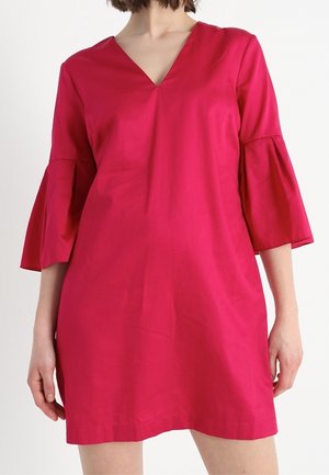 Femme portant une robe rose vif à encolure en V avec des manches trois-quarts évasées, posant devant un fond blanc uni.