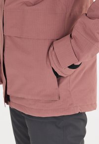 Rosa technische Jacke mit gerippter Textur, ausgestattet mit einem Reißverschluss vorne, Seitentaschen und einem verstellbaren schwarzen Klettverschluss an den Bündchen.