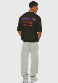 Schwarzes T-Shirt mit kurzen Ärmeln und blau-rotem Text auf dem Rücken, kombiniert mit hellgrünen Hosen und weißen Sneakers. Einfacher, lockerer Schnitt.