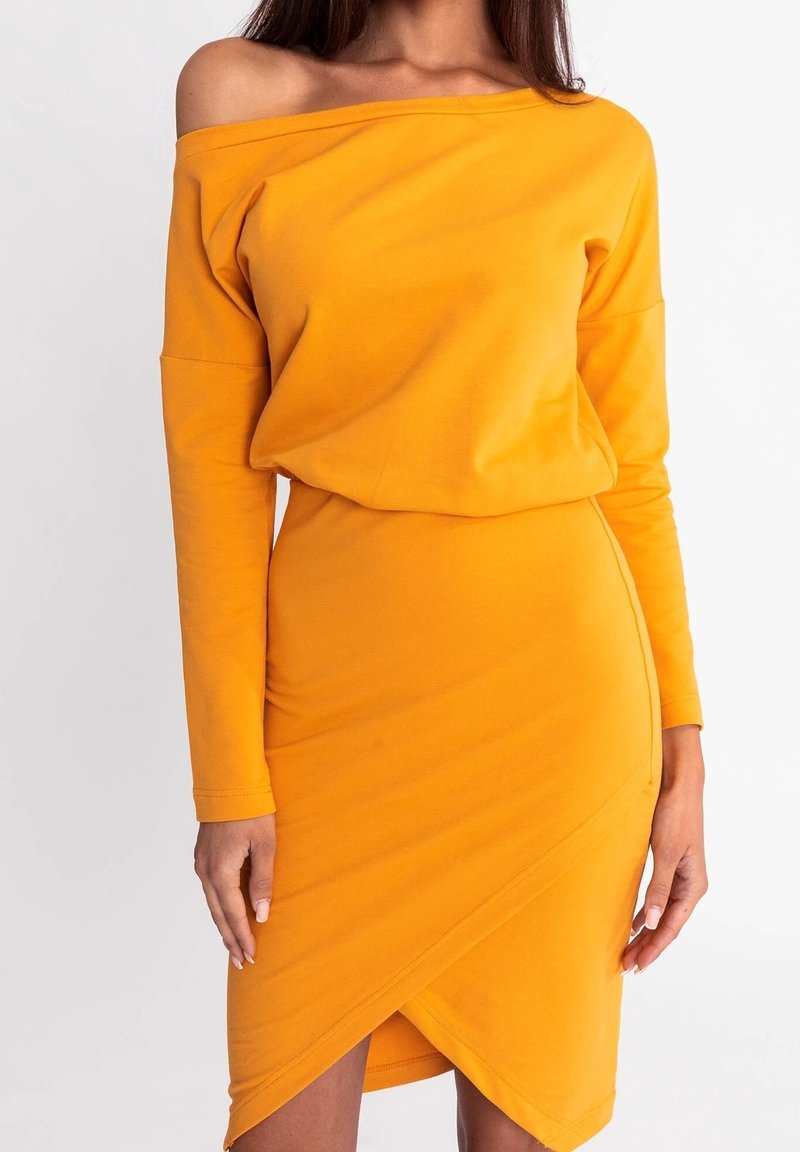 Femme portant une robe à manches longues jaune moutarde à épaules dénudées avec une jupe portefeuille asymétrique ajustée.