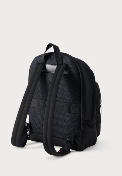 Μαύρο υφασμάτινο σακίδιο πλάτης με επενδυμένους ιμάντες ώμου, λαβή για μεταφορά από πάνω και πλαϊνή τσέπη με φερμουάρ, εμφανιζόμενο από πίσω σε λευκό φόντο.