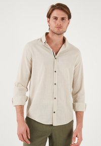Camisa de manga larga en color beige claro, hecha de tejido texturizado, con un cuello abotonado, botones de acento negros y mangas arremangadas.