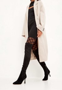 Abrigo largo beige sobre un vestido negro de canalé, combinado con botas de ante negras hasta el muslo con tacón de stiletto y medias con patrón de tablero de ajedrez.