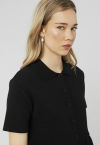 Robe en tricot noire à manches courtes, col et boutons ronds. Texture lisse et corsage ajusté, se terminant par une jupe évasée.