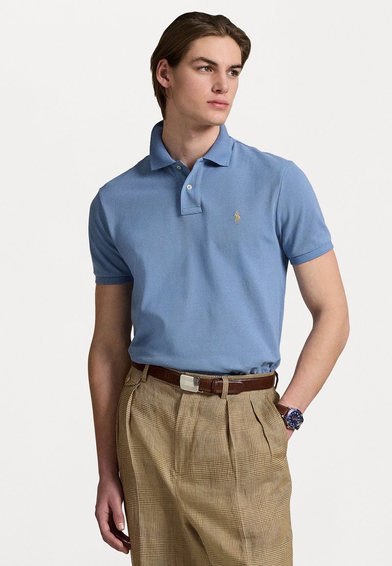 Camisa polo azul claro con un acabado texturizado y dos botones, combinada con pantalones de cuadros beige y un cinturón de cuero marrón.
