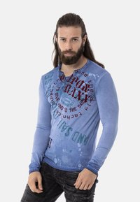 Blaues Langarmhemd mit grafischem Druck in rotem und grünem Text, das eine abgenutzte Textur und ein Rundhals-Design aufweist.