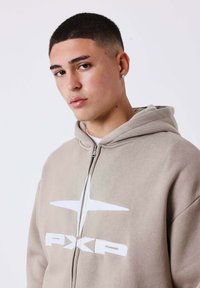 Beige zip-up hoodie met een wit logo, voorzien van een capuchon en een relaxte pasvorm. Soepele textuur met zichtbare stitching en een voorkant met rits.