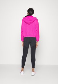 Haut à capuche texturé rose, associé à des leggings noirs avec des panneaux contrastés et des chaussures de sport blanches.