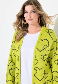 Cardigan lavorato a maglia di un vivace verde brillante con motivi a cuore neri. Design aperto davanti abbinato a una maglietta bianca. Maniche lunghe e texture morbida.