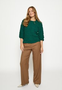 Zelený textúrovaný top s okrúhlym výstrihom a rukávmi do troch štvrtín, v kombinácii s hnedými širokými nohavicami a krémovými špicatými topánkami.