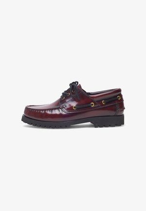 Scarpa da barca in pelle bordeaux con punta arrotondata, lacci neri, occhielli dorati e suola in gomma nera texturizzata con motivo a ondulazione.
