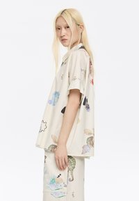 Camicia a maniche corte, leggero beige, con stampe floreali e animali colorate. Dotata di colletto con bottoni e vestibilità ampia e rilassata.