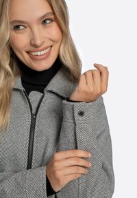 Graue Fischgrätjacke mit Reißverschluss vorne, großem Kragen und Knopfdetails. Schwarzer, gerippter Rollkragenpullover darunter. Glatte Textur.