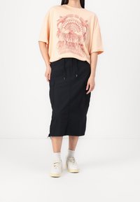 Camiseta de gran tamaño en color durazno con estampado gráfico, combinada con una falda midi negra con cintura ajustable y bolsillos laterales, llevada con zapatillas blancas.