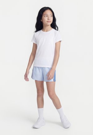 Κορίτσι με μακριά μαύρα μαλλιά στέκεται κοιτάζοντας δεξιά, φορώντας λευκό T-shirt, ανοιχτό μπλε σορτς, λευκές κάλτσες και λευκά αθλητικά παπούτσια σε απλό φόντο.