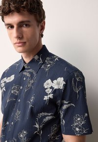 Kurzärmeliges marineblaues Hemd mit strukturierter Oberfläche, versehen mit weißen Blumenmustern, einem Button-Down-Kragen und Vorderknöpfen.
